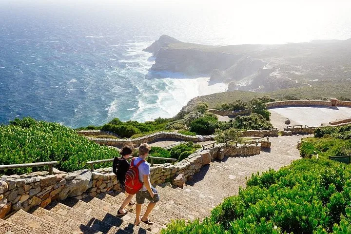 Zwei Personen gehen eine Steintreppe hinunter und blicken auf eine Küstenlandschaft mit Meer.